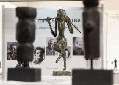 Бронзана скулптура „Сатир“ Владете Петрића у крупном плану. Стилизована, издужена фигура митолошког бића приказана је у динамичном ставу, на једној нози, са рукама које држе штап или фрулу иза врата. У позадини се види пано изложбе „Мајстори вајарства“ са портретима уметника, док су у предњем плану две замагљене тамне скулптуре које уоквирују централни експонат.